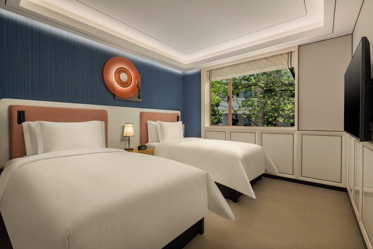 Ein Hotelzimmer mit zwei Betten, einem großen Fenster mit Blick auf Bäume, einem Flachbildfernseher und einer Wanduhr.