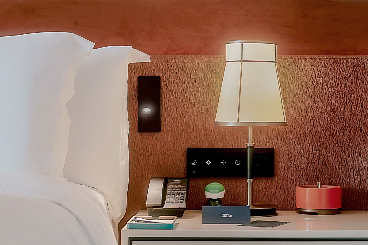 Ein Hotelzimmer mit einem Bett, einem Nachttisch mit Lampe, einem Telefon, einer Minibar und einer wandmontierten Steuerung.