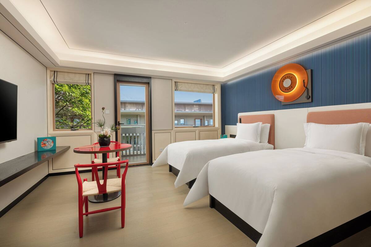 Habitación de hotel con dos camas, televisión, mesa de comedor con sillas y balcón con vista al edificio.