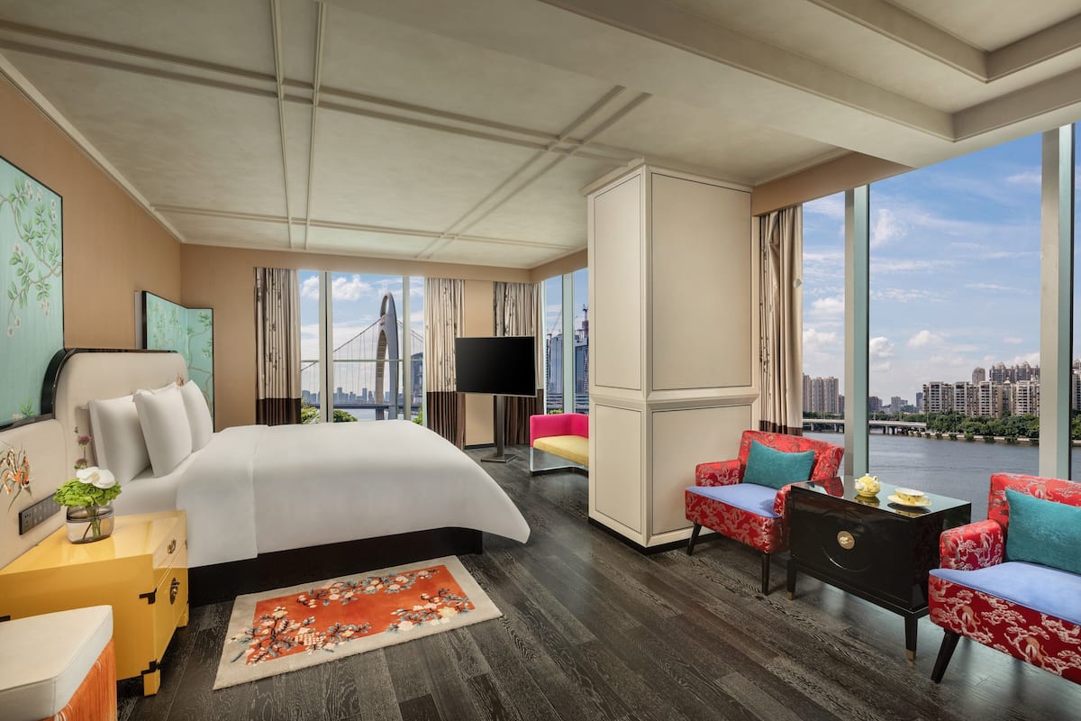 Una habitación de hotel moderna con una cama grande, televisión, escritorio, silla y vistas a un paisaje urbano a través de grandes ventanales.