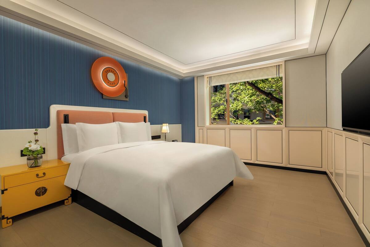 Habitación de hotel con cama, mesita de noche, televisor y ventana con vistas al verdor.
