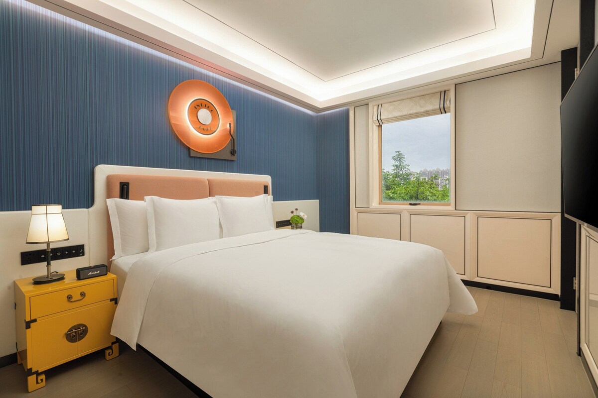 Ein Hotelzimmer mit einem großen Bett, einem Nachttisch mit Lampe, einer Wanduhr und einem Fenster mit Blick ins Grüne.
