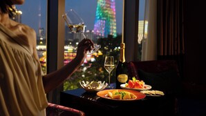 Une personne tenant un verre de vin, assise à une table devant une bouteille de champagne, une assiette de nourriture et un saladier de fruits. On aperçoit, à travers la fenêtre, un paysage urbain avec une tour illuminée.