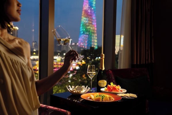 Une personne tenant un verre de vin, assise à une table devant une bouteille de champagne, une assiette de nourriture et un saladier de fruits. On aperçoit, à travers la fenêtre, un paysage urbain avec une tour illuminée.