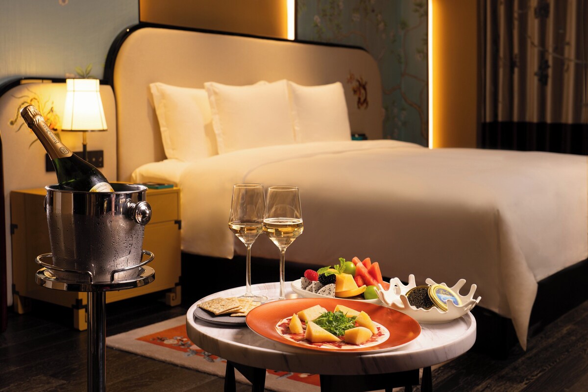 Une chambre d’hôtel avec un lit, une table de chevet sur laquelle se trouve une bouteille de champagne, deux verres de vin et une assiette de fruits.