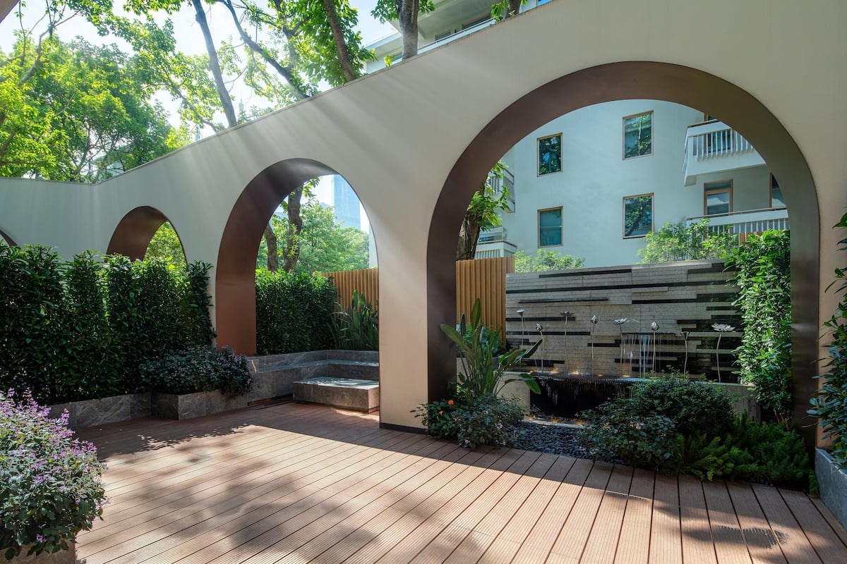 Una terraza de madera con arcos, rodeada de follaje y un elemento acuático.