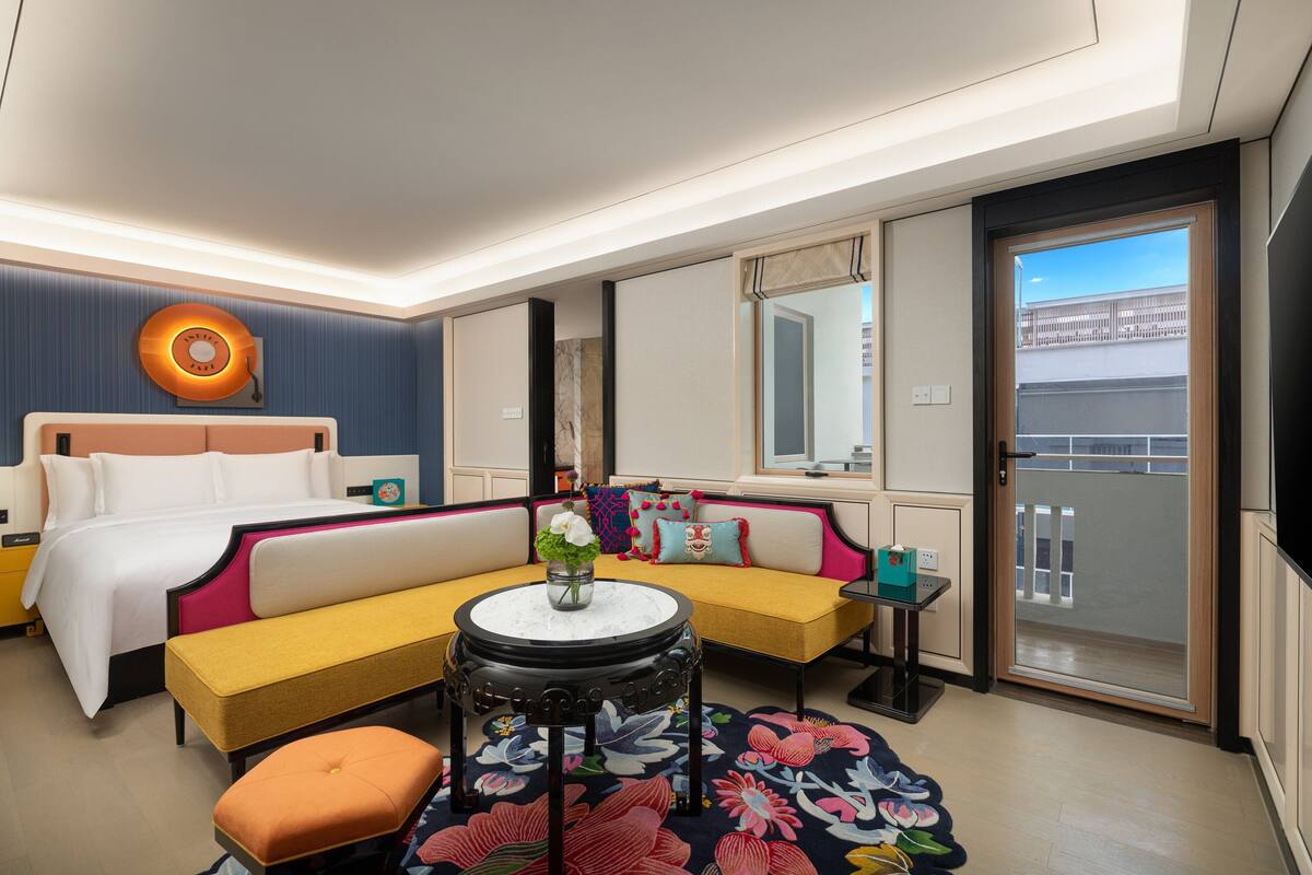 Una habitación de hotel con una cama, un sofá, una mesita con un florero, un banco y una alfombra colorida.