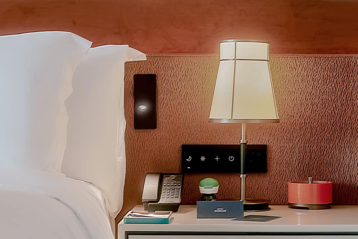 Habitación de hotel con cama, mesita de noche con lámpara, teléfono, minibar y panel de control en la pared.
