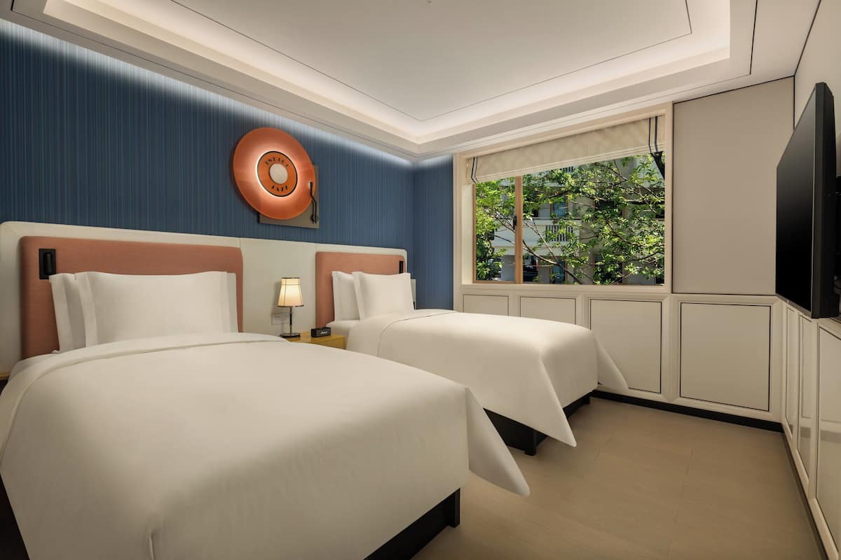 Habitación de hotel con dos camas, un ventanal con vistas a árboles, un televisor de pantalla plana y un reloj de pared.