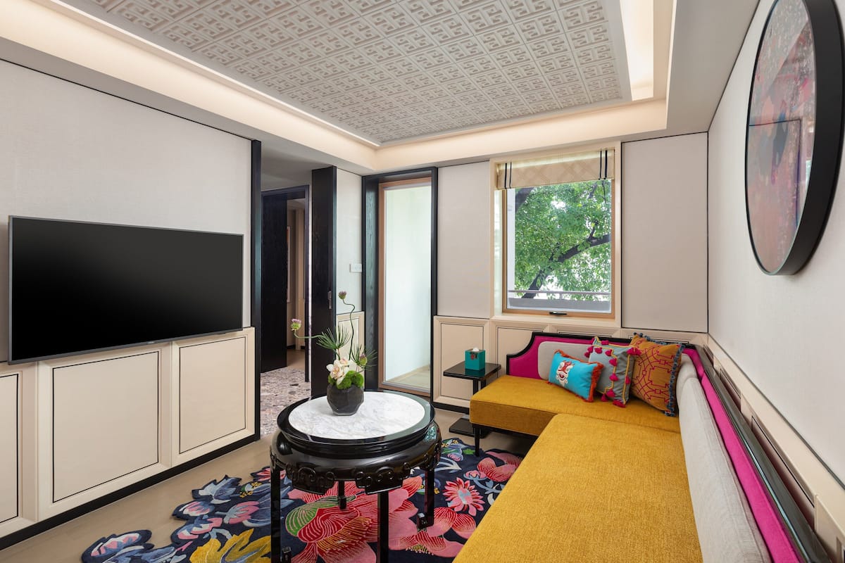 Una sala de estar moderna con un televisor de pantalla plana, una mesa redonda con un jarrón, un sofá con cojines de colores y un ventanal con vistas a árboles.