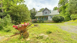 A house with a porch, surrounded by lush greenery and a wheelbarrow with flowers. - Pet-Friendly Smoky Mountain Getaway w/ Fire Pit! (Clyde)
