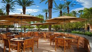 An outdoor patio area with wooden tables and chairs, large umbrellas, and a pool in the background.