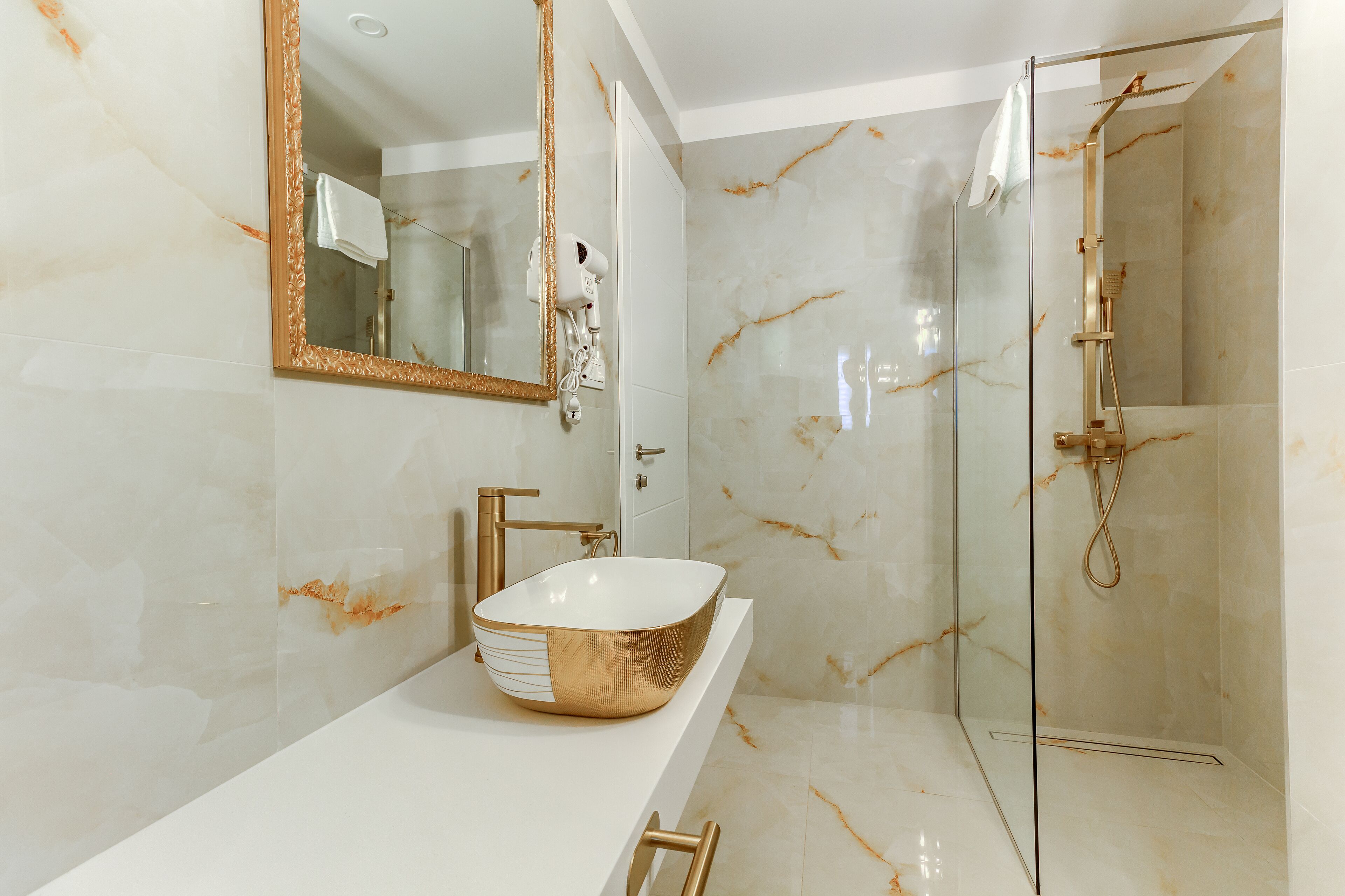 Un baño con un espejo grande, un lavabo tipo recipiente dorado, cabezal de ducha de mano y paredes de mármol.