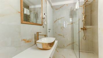 Un baño con un espejo grande, un lavabo tipo recipiente dorado, cabezal de ducha de mano y paredes de mármol.