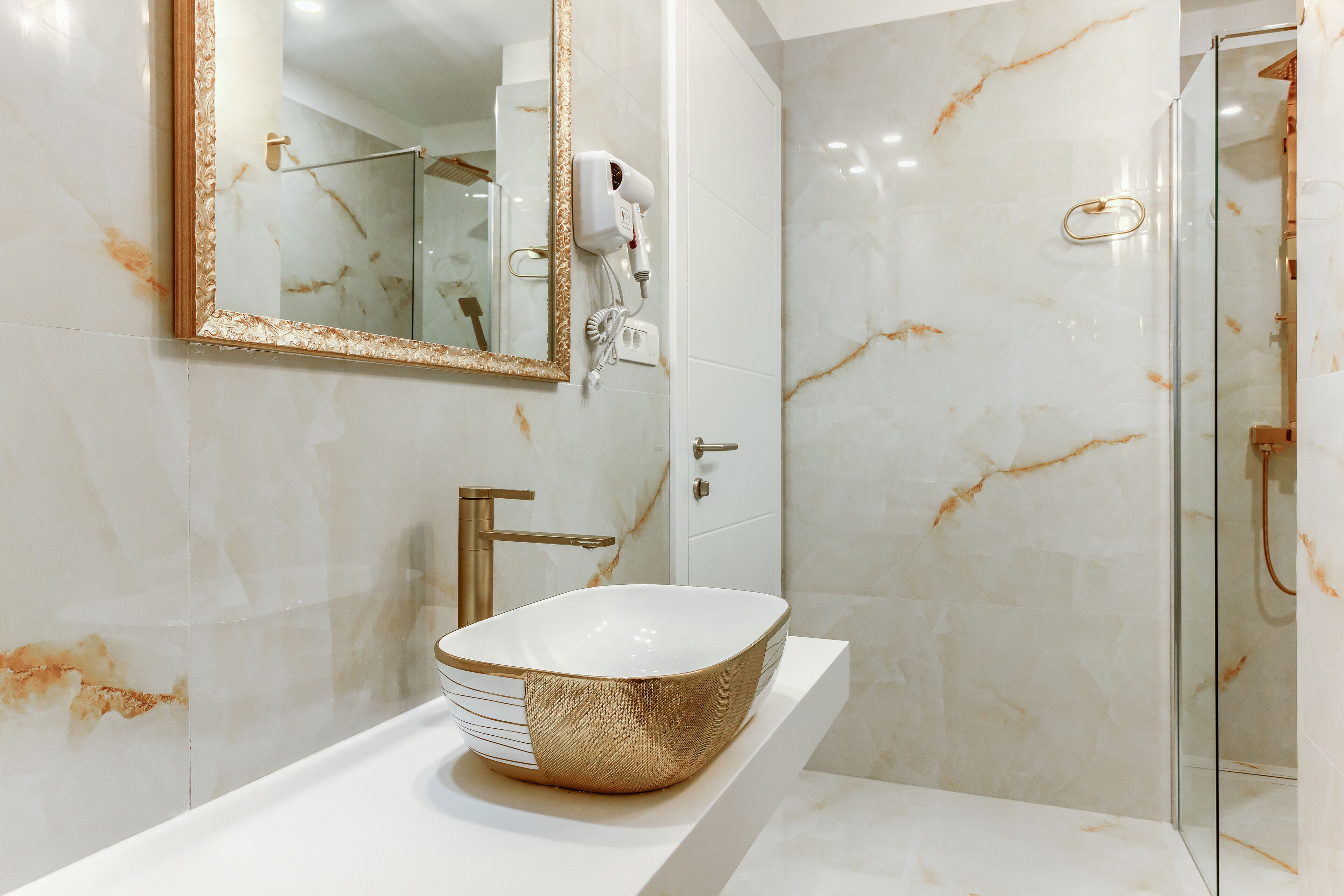 A modern bathroom with a large mirror, a gold-colored vessel sink, a handheld hair dryer, and a glass shower enclosure.