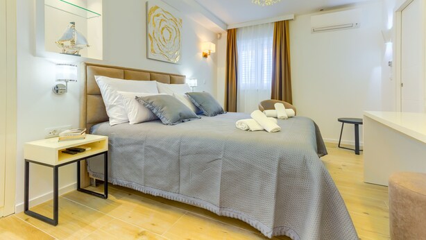 A modern hotel room with a large bed, a bedside table with a lamp, a small desk, and a chair. - Irini Luxury Rooms (Split)