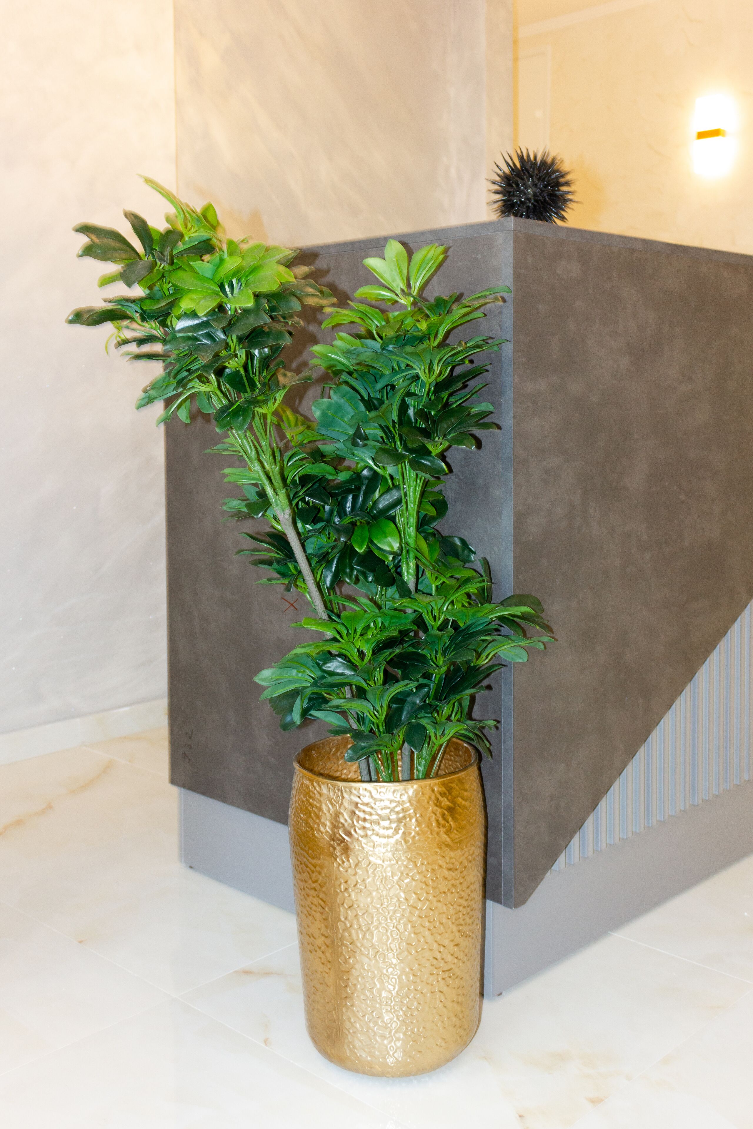 Una planta en maceta con follaje verde en un jarrón dorado texturizado, sobre un piso de baldosas frente a una pared gris con una lámpara.