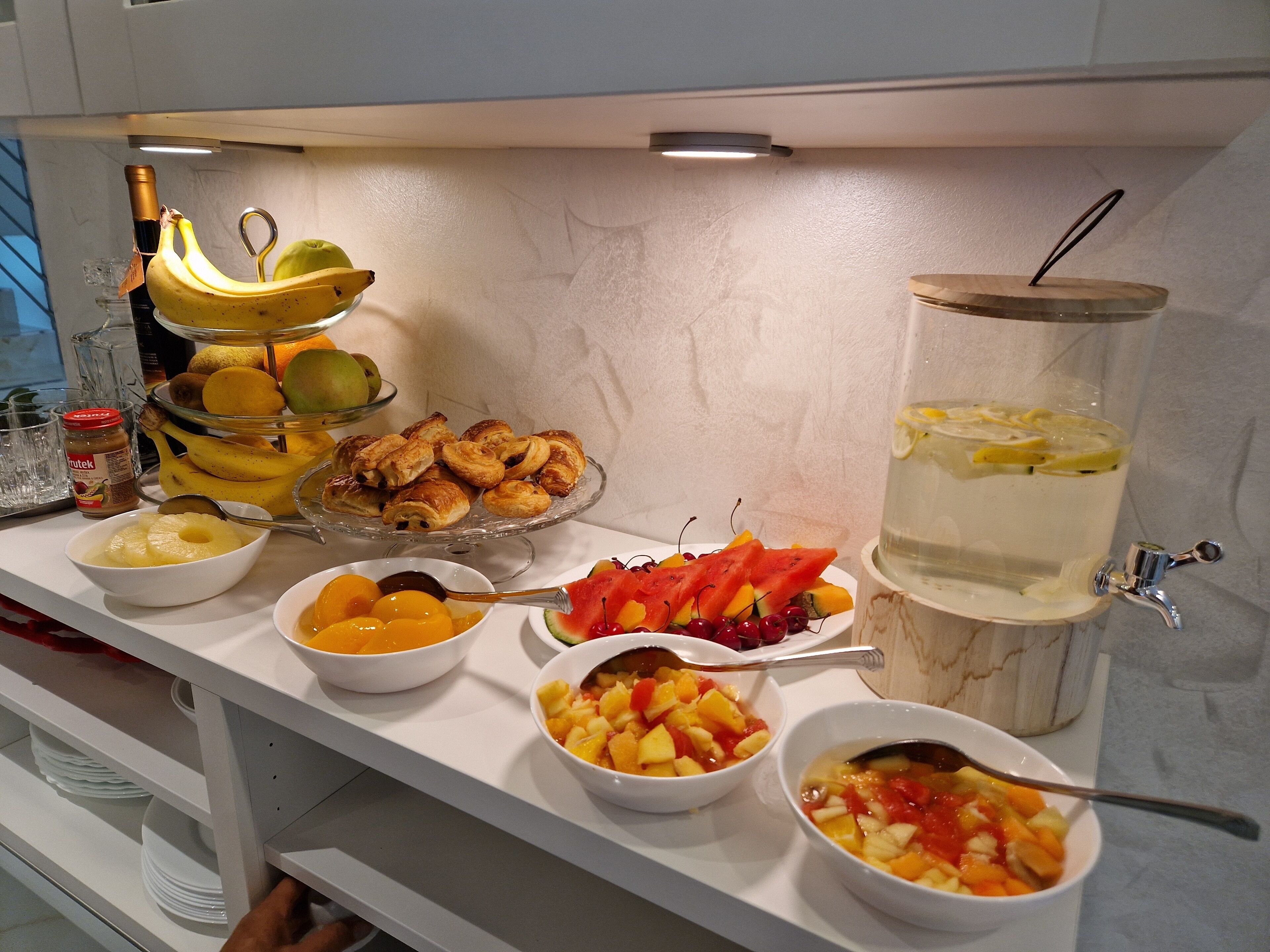 Une table de buffet proposant une variété de fruits, de pâtisseries et un distributeur de boissons.