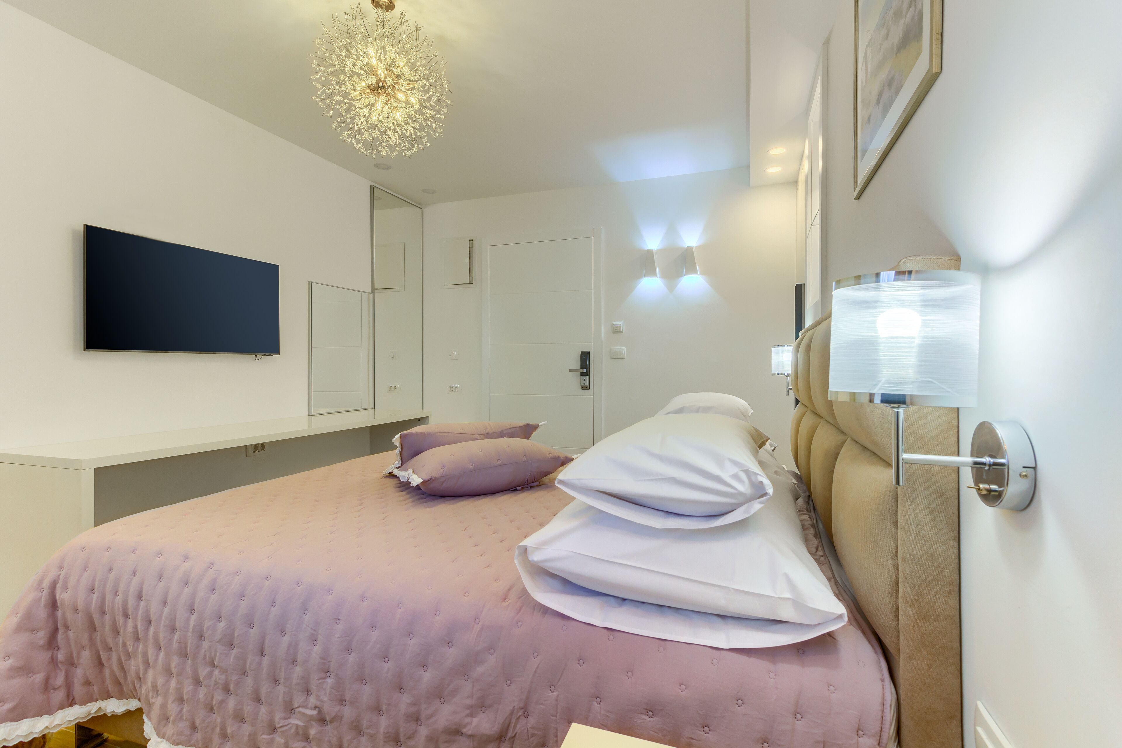 Un dormitorio con cama, almohadas, un televisor de pantalla plana, una lámpara de pared y una puerta.