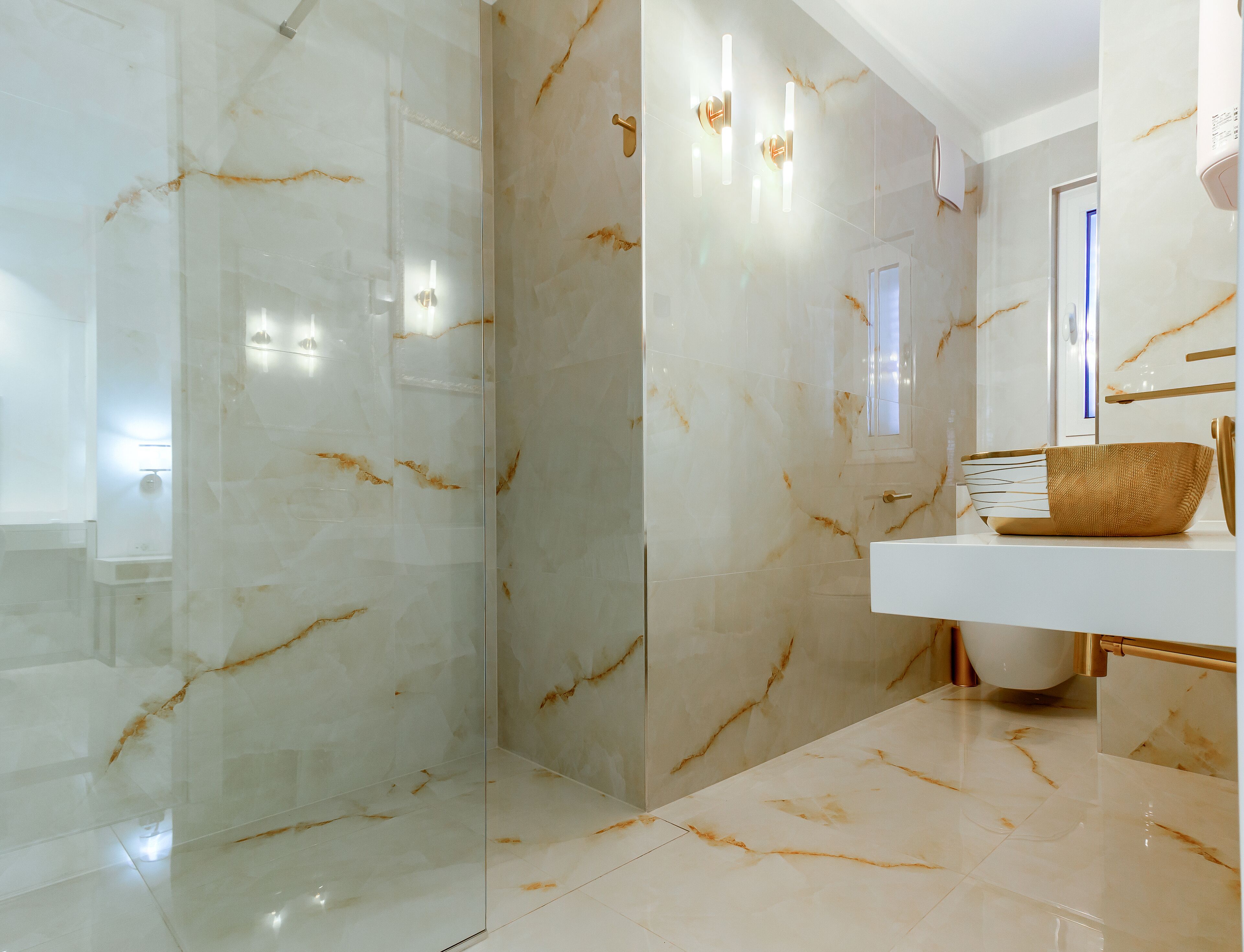 Une salle de bain moderne avec une douche à l’italienne en verre, un meuble-lavabo blanc avec un vasque dorée et des murs en marbre.