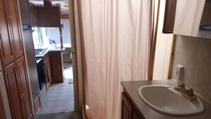 Ein Badezimmer mit Waschbecken, Holzausstattung, einer Duschvorhang und einem sichtbaren Küchenbereich im Hintergrund.
