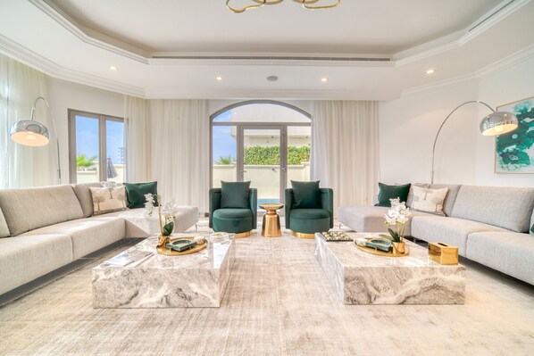 A spacious living room with a large sofa, two armchairs, and a coffee table. There is a large window with curtains, a door leading outside, and a ceiling light fixture.