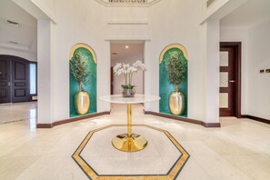 A lobby with a central round table, two large vases with plants, and a geometric patterned rug.