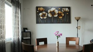 Et spiseområde med et rundt træbord, fire stole, en vase med blomster, et billede over bordet, en lampe og en reol med en telefon og en lille genstand.