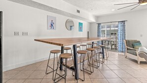 A modern kitchen with a long wooden bar, bar stools, and a dining area with a white sofa and patterned curtains. - bees knees (Myrtle Beach)