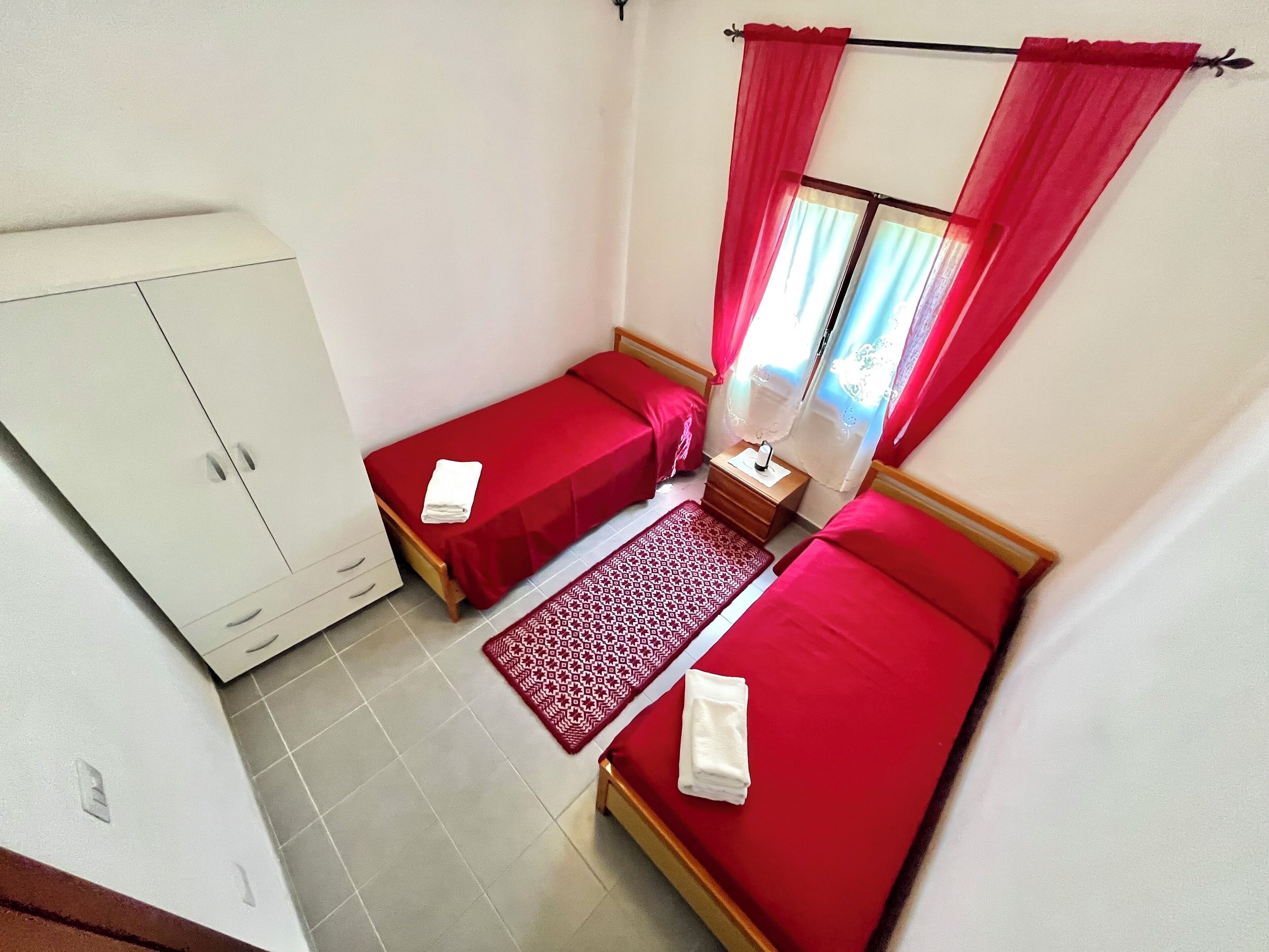 Une petite chambre propre, avec deux lits simples, une armoire, une table de chevet avec une lampe et un tapis à motifs.