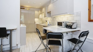 Une cuisine moderne avec des meubles blancs, un four à micro-ondes, un grille-pain et un évier. On y trouve trois chaises noires et un comptoir blanc.