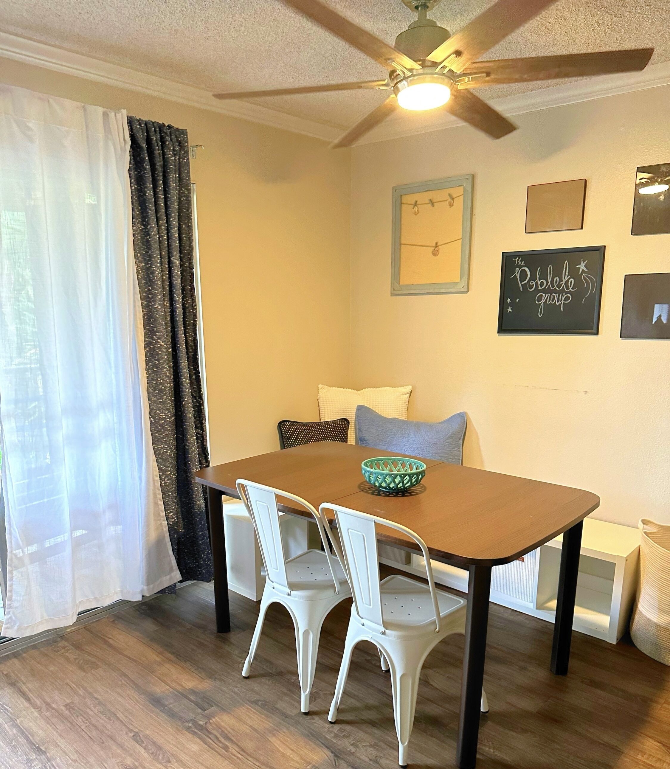 A dining area with a wooden table, white chairs, a ceiling fan, and a wall with framed pictures and a sign.