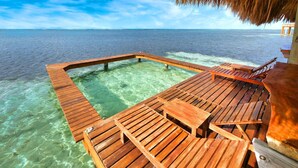 Una terraza de madera con piscina, sillas de descanso y una estructura con techo de paja.