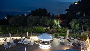 En terrasse med liggestole og en kiosk i midten, med udsigt over et kystområde med træer og en by i det fjerne.
