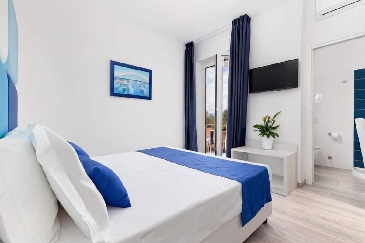 Habitación de hotel con cama, televisor montado en la pared, una mesita con planta y ventana con cortinas.