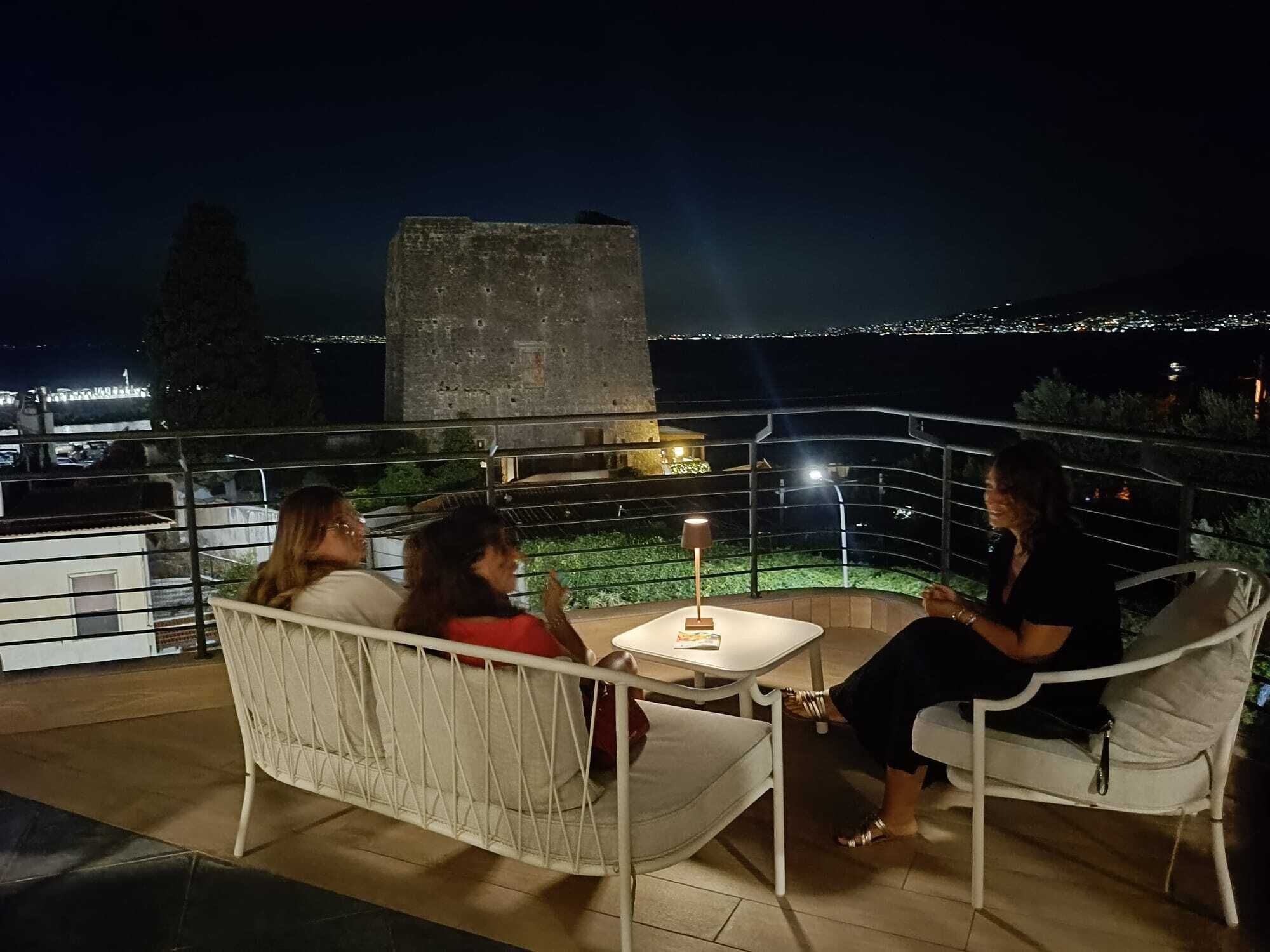 Una terraza con zona de estar exterior blanca, una mesa con lámpara y vistas a un paisaje urbano nocturno.