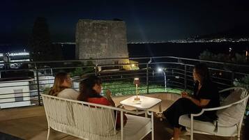 Una terraza con zona de estar exterior blanca, una mesa con lámpara y vistas a un paisaje urbano nocturno.