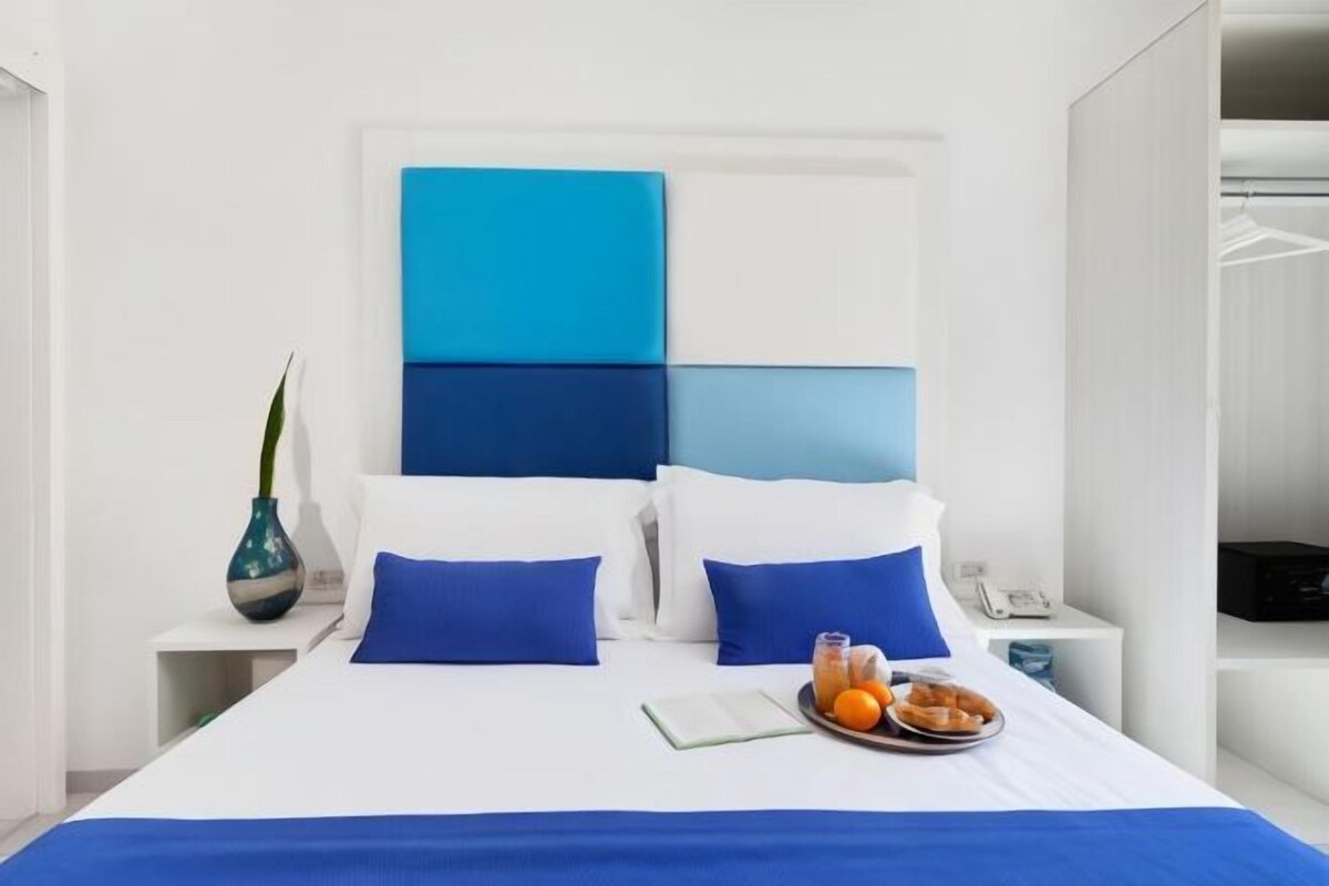 A hotel room with a bed, a bedside table with a vase, and a tray with fruits and a book.