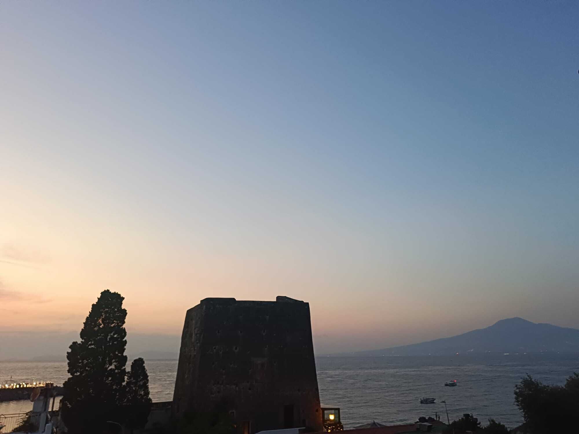 A coastal view with a historic stone building, a clear sky, and a distant mountain.