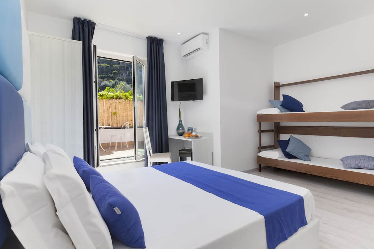 Una habitación de hotel moderna con una cama grande, una cama litera, un escritorio con televisor y un puerta corrediza de vidrio a un balcón.