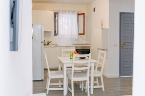 Eine kleine Küche mit weißen Schränken, einem Esstisch mit Stühlen, einem Kühlschrank und einem kleinen Fenster mit Vorhang.