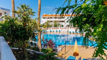 Una piscina con lettini e ombrelloni, circondata da palme e da un edificio con tetto in tegole.
