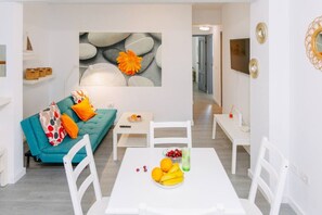 Un salon moderne avec un canapé turquoise, des chaises blanches, une table blanche ornée de fruits et une œuvre d’art murale en pierre.