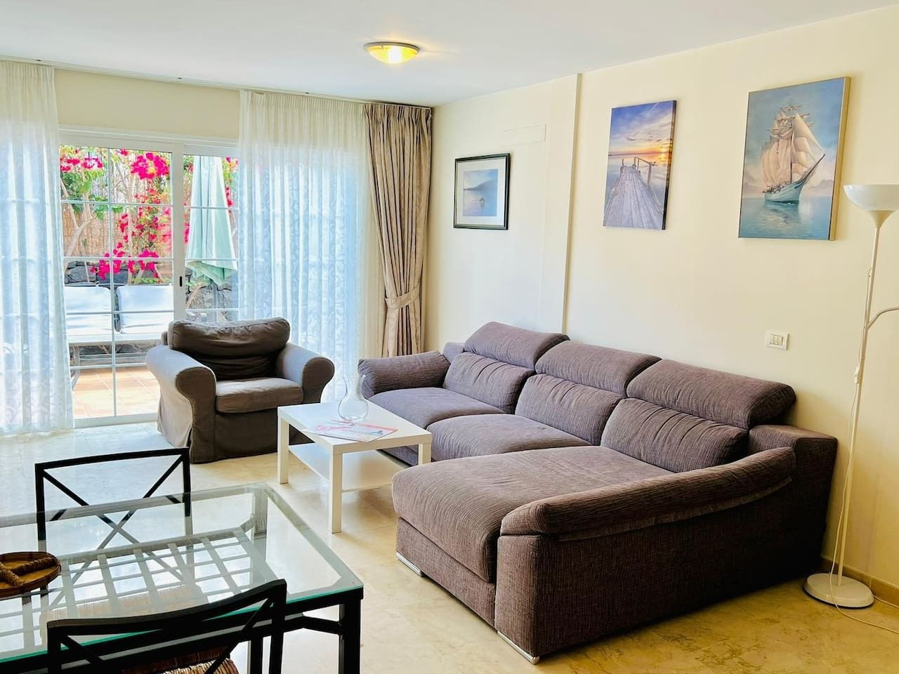 A living room with a sofa, a coffee table, and a dining table with chairs. There are two paintings on the wall and a window with curtains.
