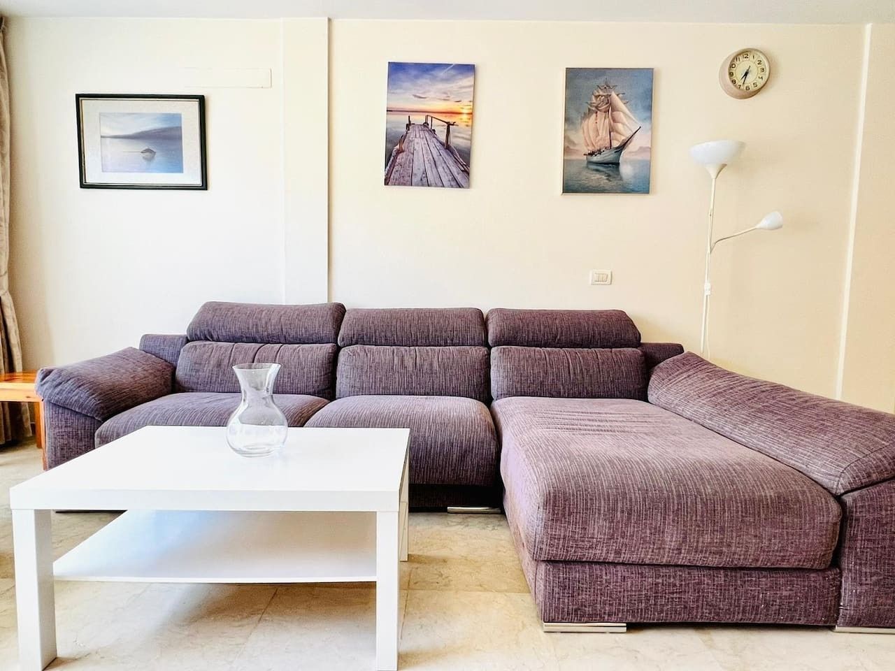 Un salón con un sofá modular, una mesa de centro blanca y obras de arte, incluyendo una escena de atardecer y un velero.