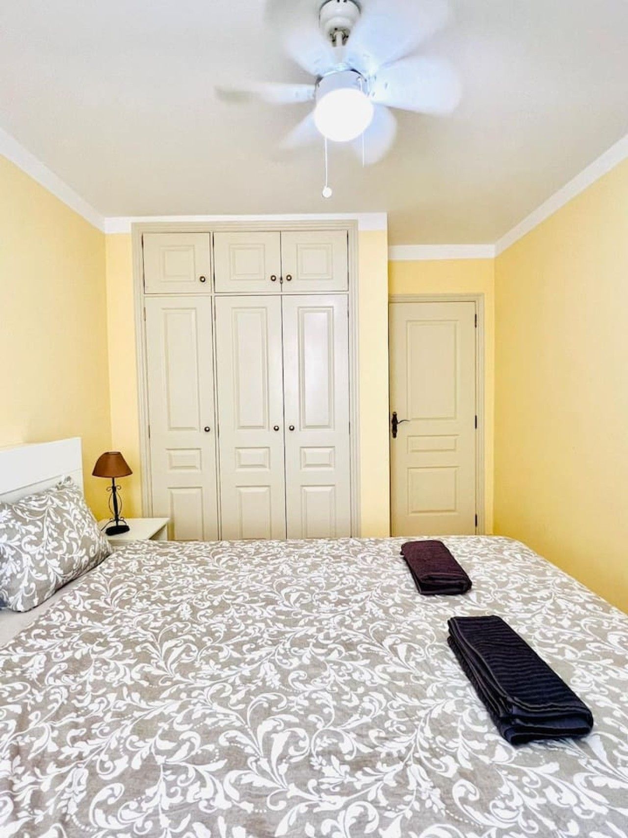 A bedroom with a bed, a ceiling fan, a nightstand, a lamp, and a wardrobe.