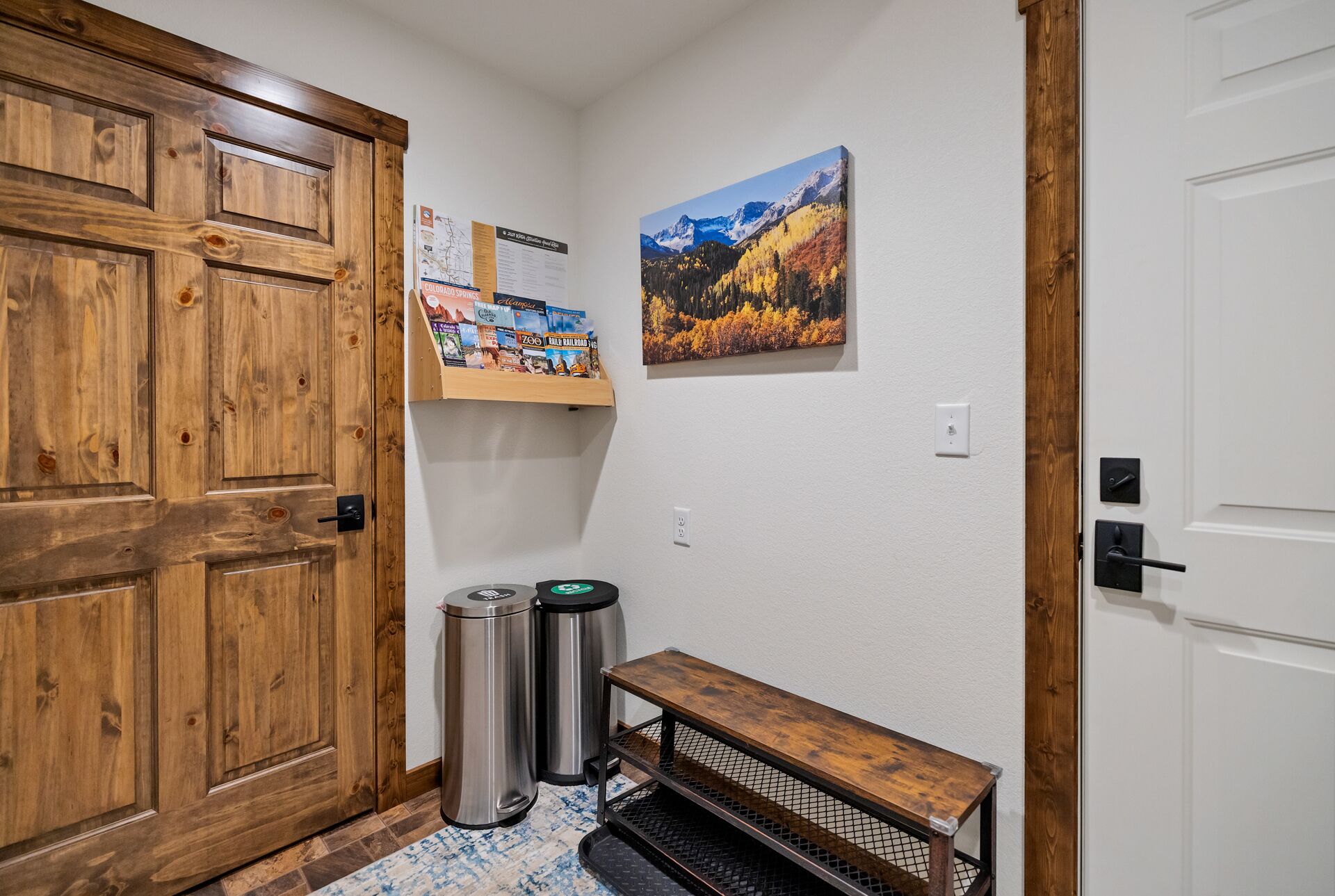 A hallway with a wooden door, a bench, trash bins, a shelf with travel brochures, and a framed landscape painting.
