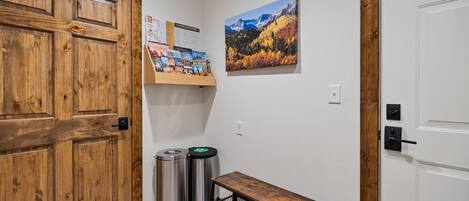 A hallway with a wooden door, a bench, trash bins, a shelf with travel brochures, and a framed landscape painting.