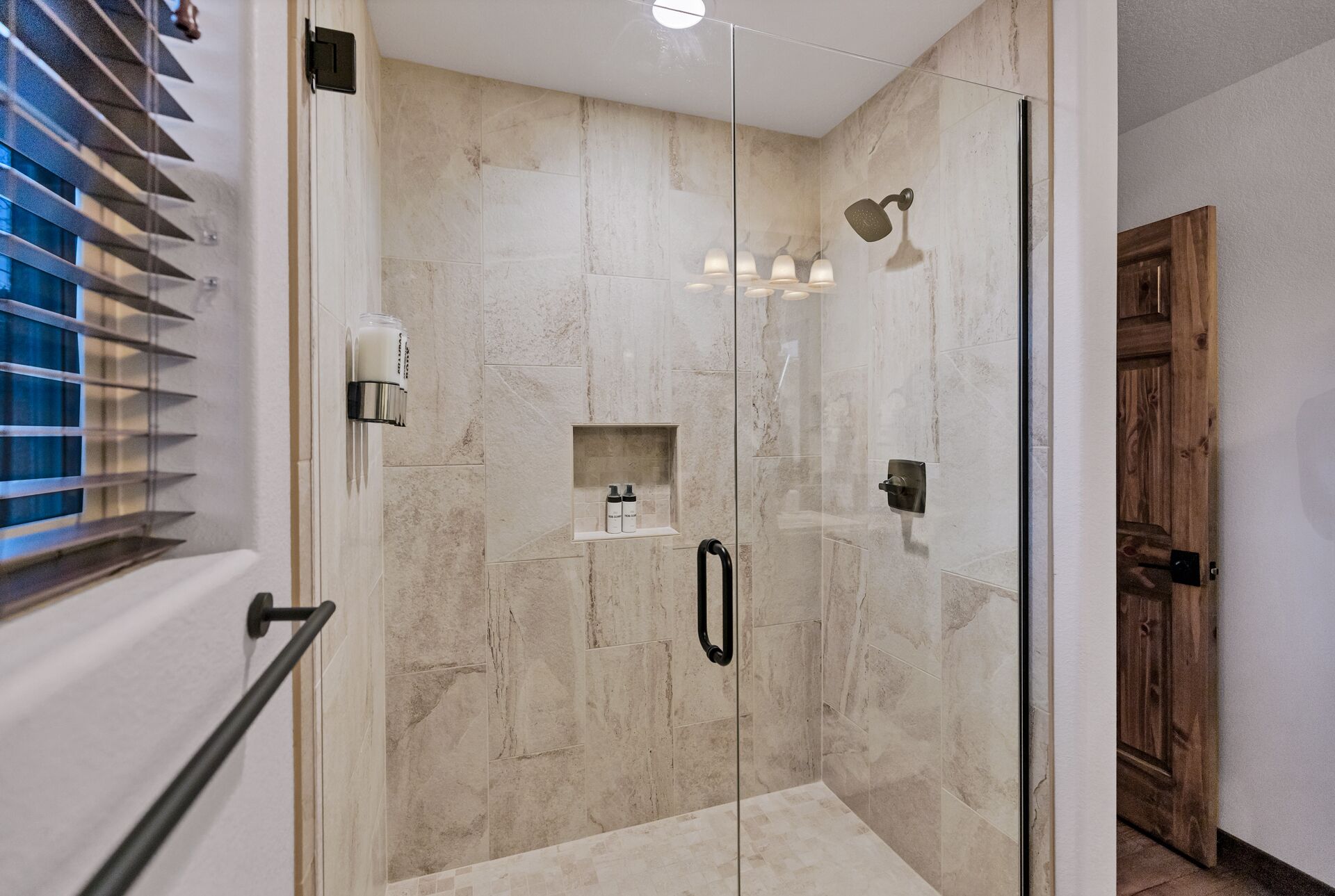 Une douche fermée par des parois de verre, avec un mur en marbre, une étagère pour les articles de toilette et une porte en bois.