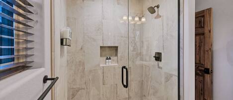 Une douche fermée par des parois de verre, avec un mur en marbre, une étagère pour les articles de toilette et une porte en bois.